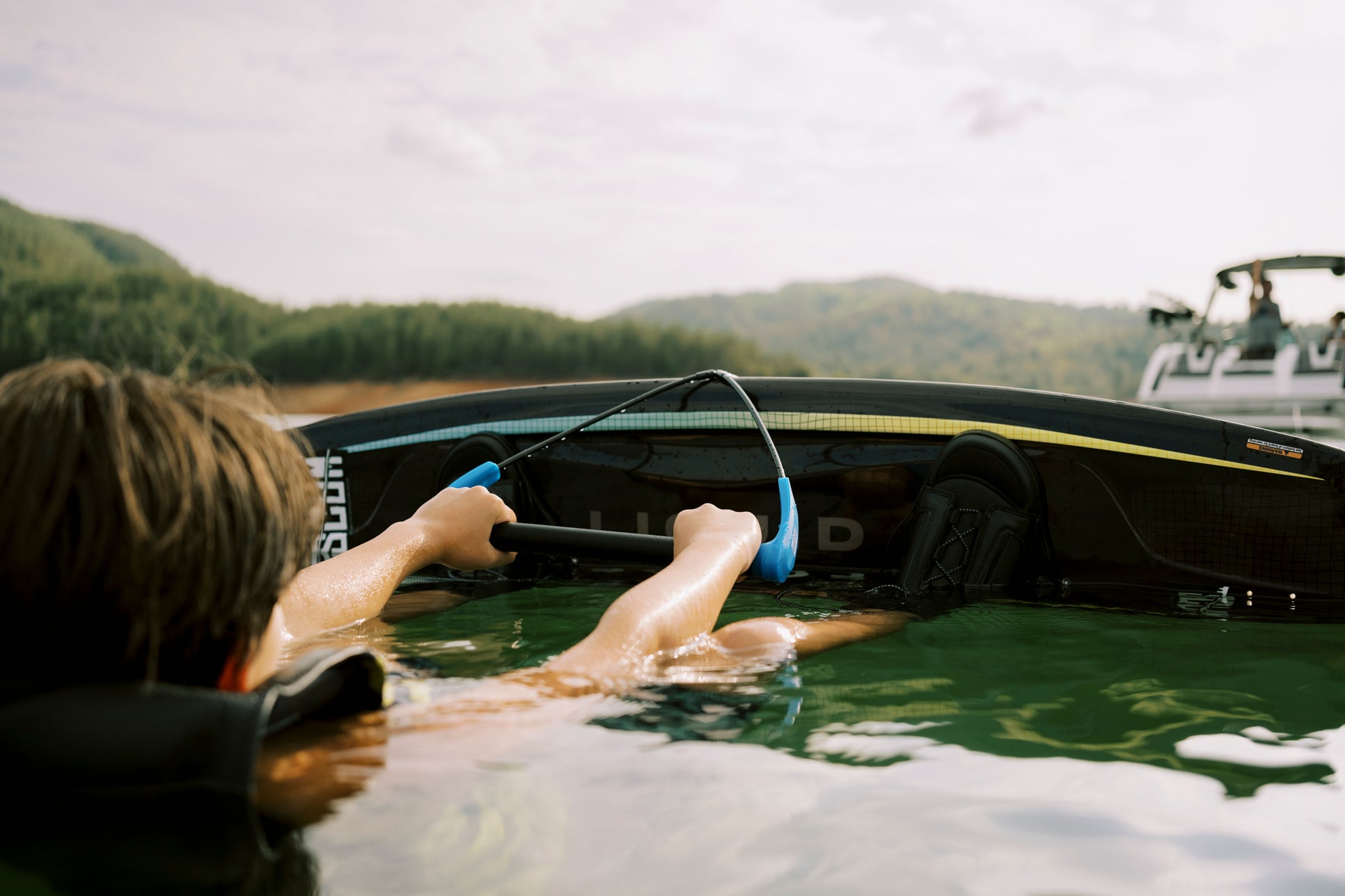 Person floating in water holding a wakeboard handle, boat in the background, surrounded by hills.