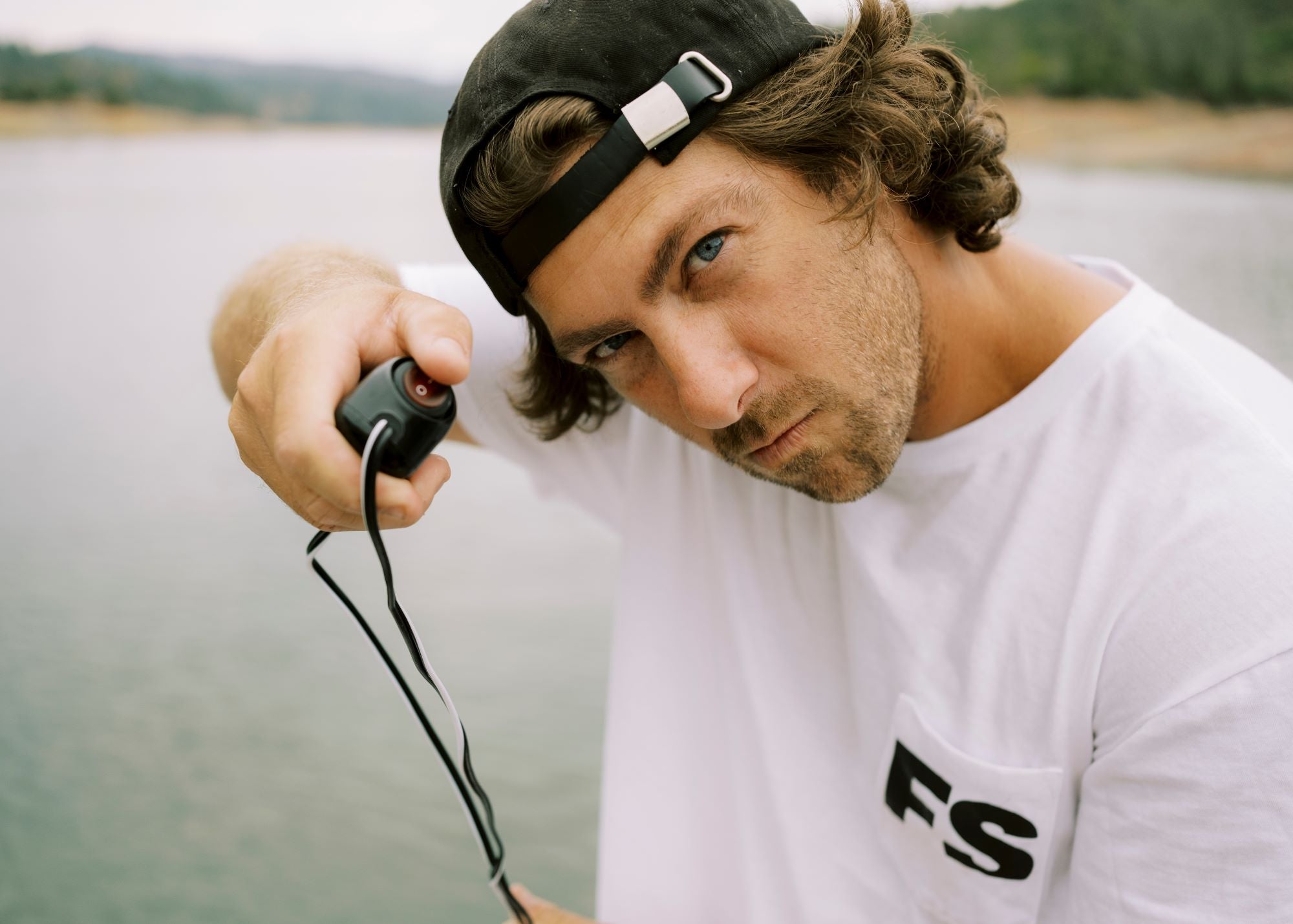 Man in a cap holds a remote by a lake, wearing a white shirt with FS logo.