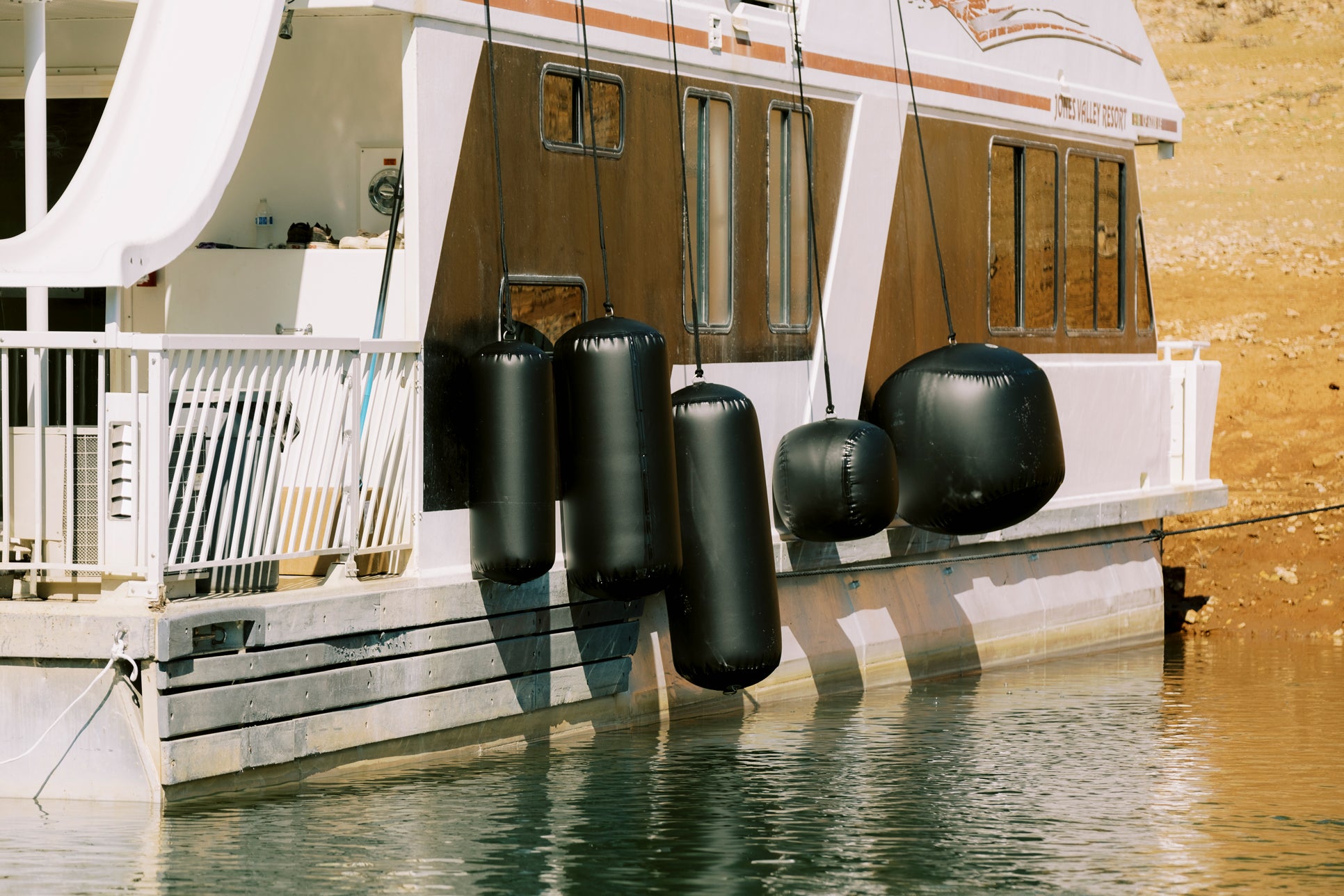 A houseboat with black fenders floats on calm water near a sandy shore.