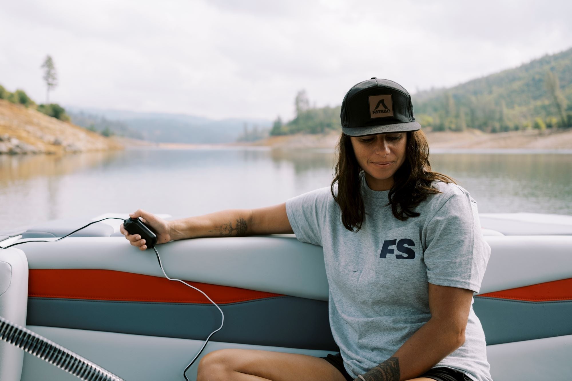 Person in a cap and t-shirt holding a device, sitting on a boat with a scenic lake and hills in the background.