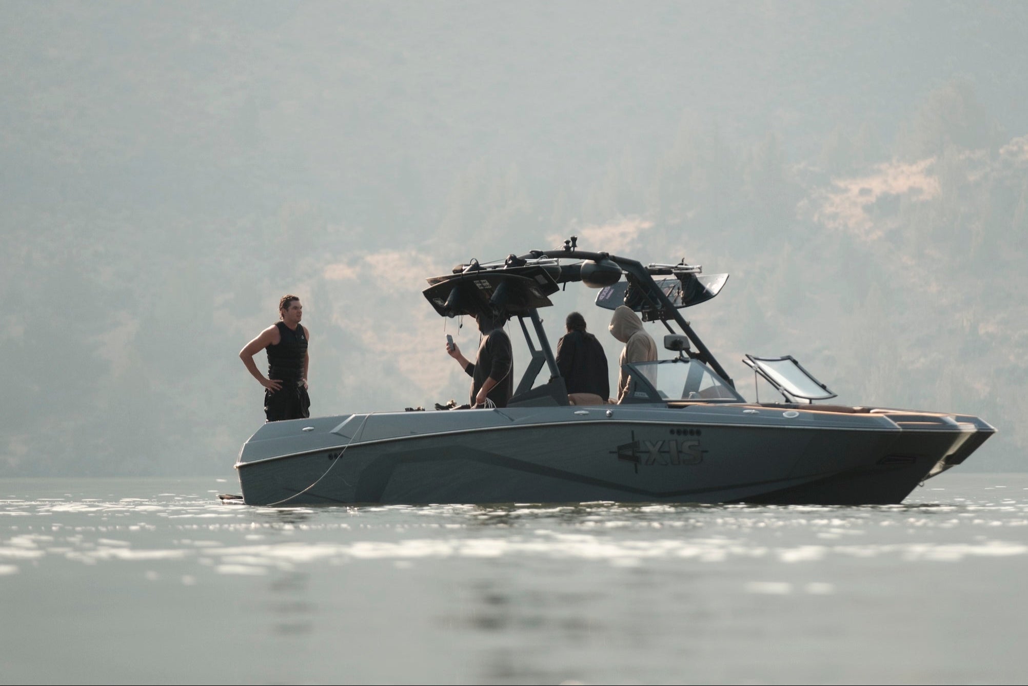 Rider stands beside boat on still water while crew records video onboard.