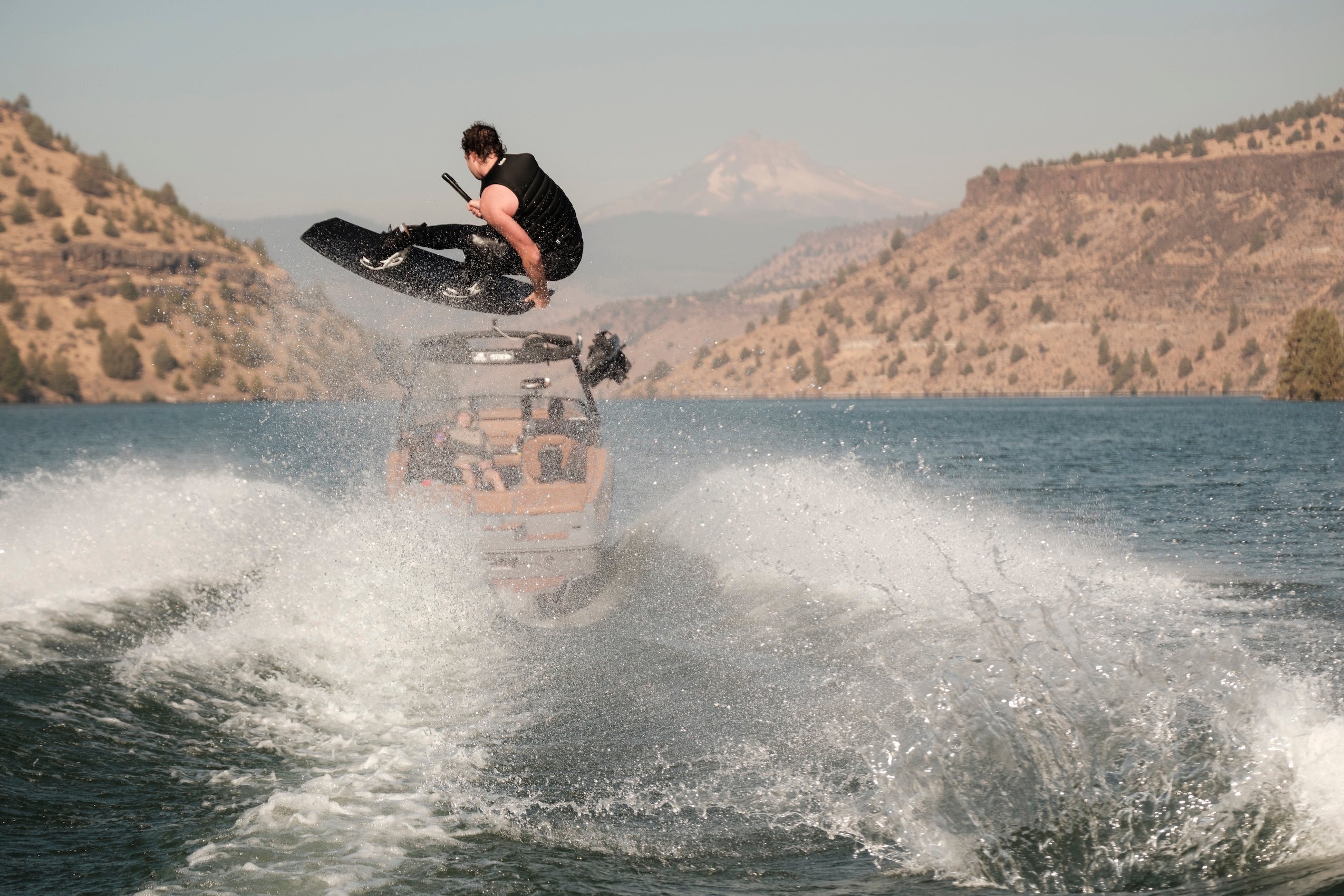 Wakeboarder in black vest grabs the board midair with Mt. Hood in the background.