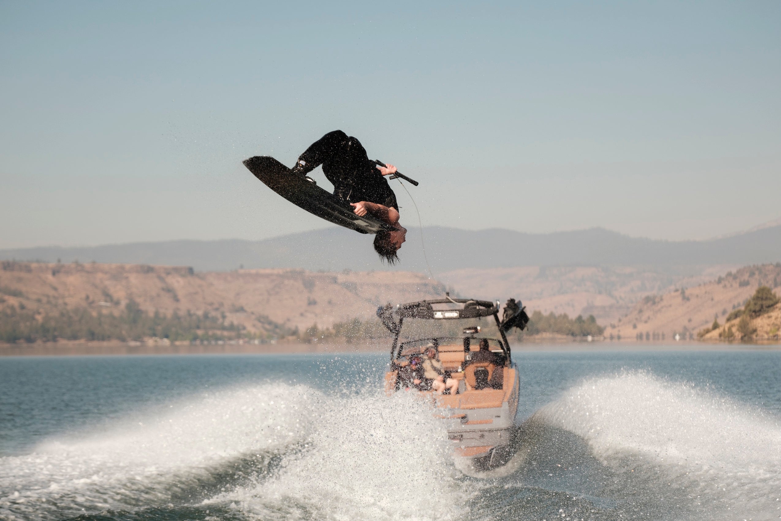 Wakeboarder performs a backflip behind the boat with dry hills in the background.
