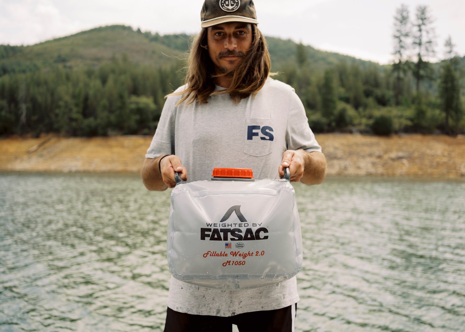 Person holding a FatSac bag by a lake with hills and trees in the background.