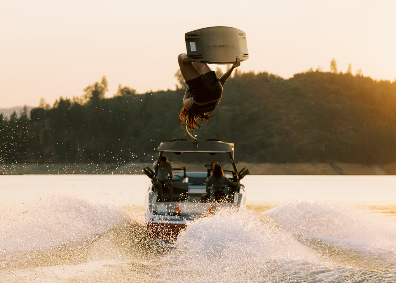 Person wakesurfing, mid-flip, at sunset with a boat in the background.