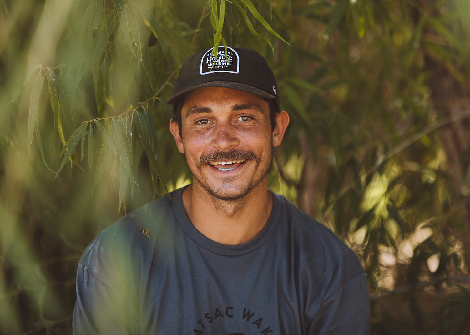 Smiling man in a black cap and gray shirt, surrounded by green foliage.