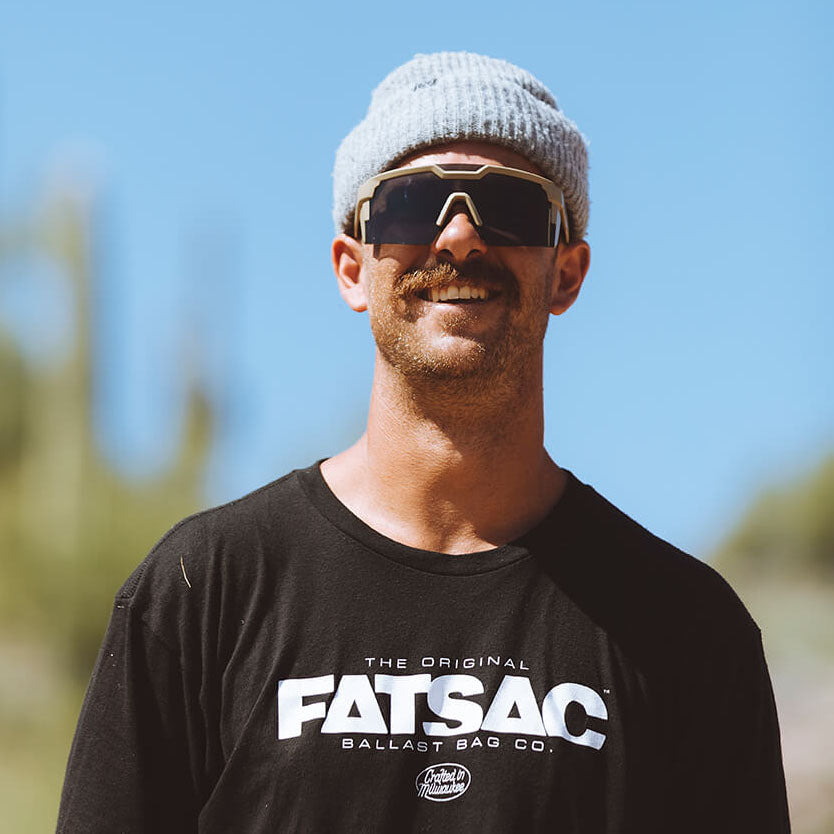 Man in a beanie and sunglasses smiling, wearing a black t-shirt outdoors.