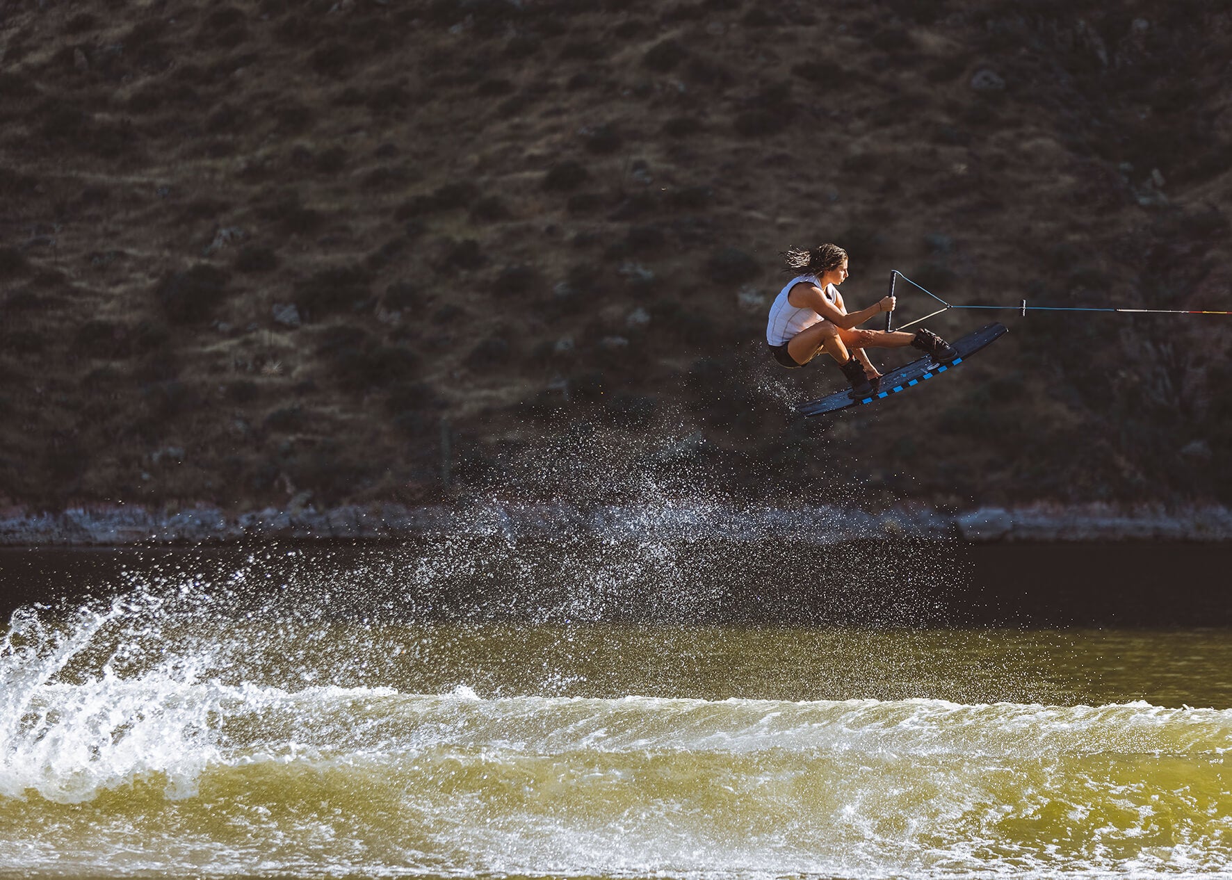 Person wakeboarding mid-air over sunlit water with a mountainous backdrop.