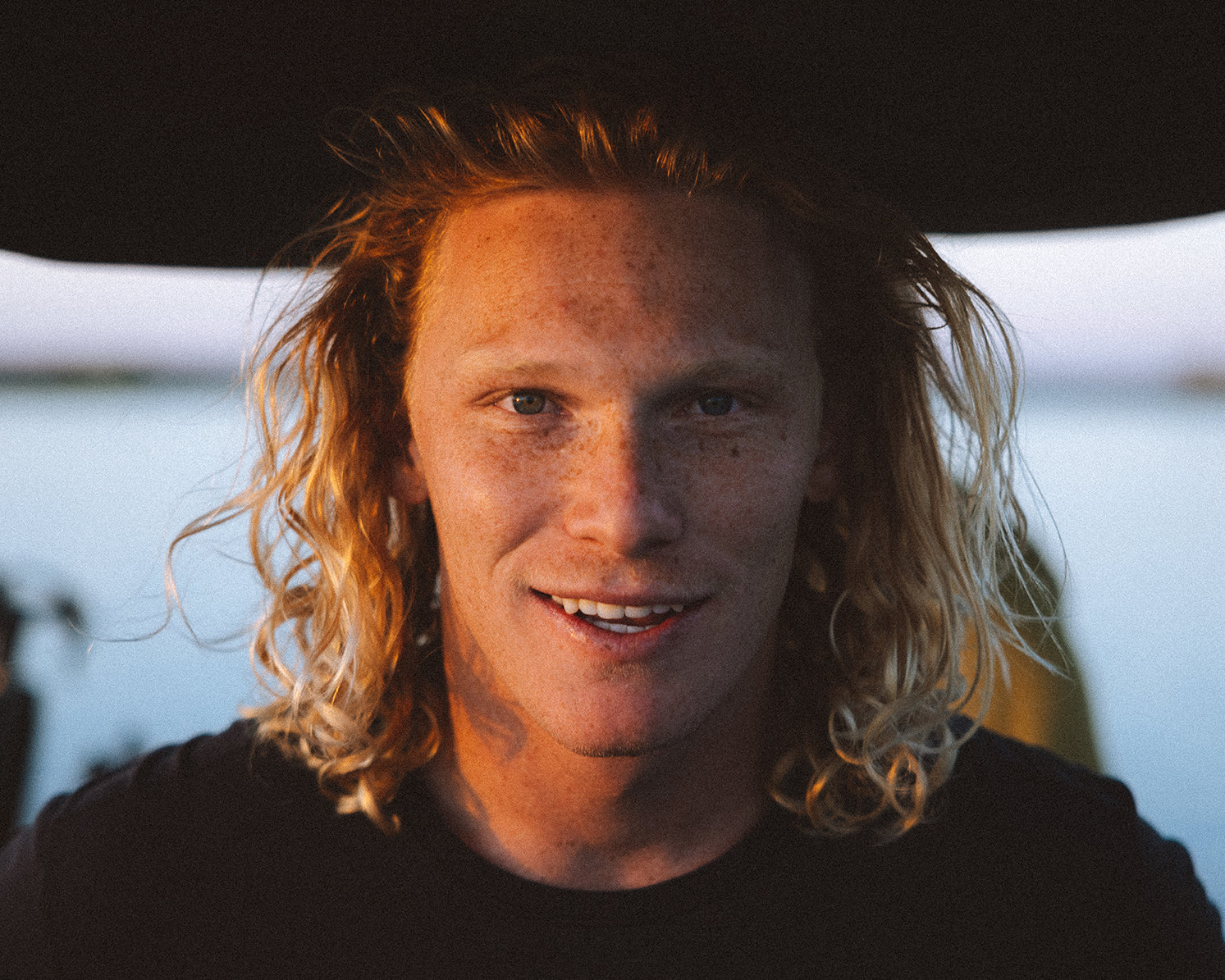 Person with long curly hair smiling outdoors near water.