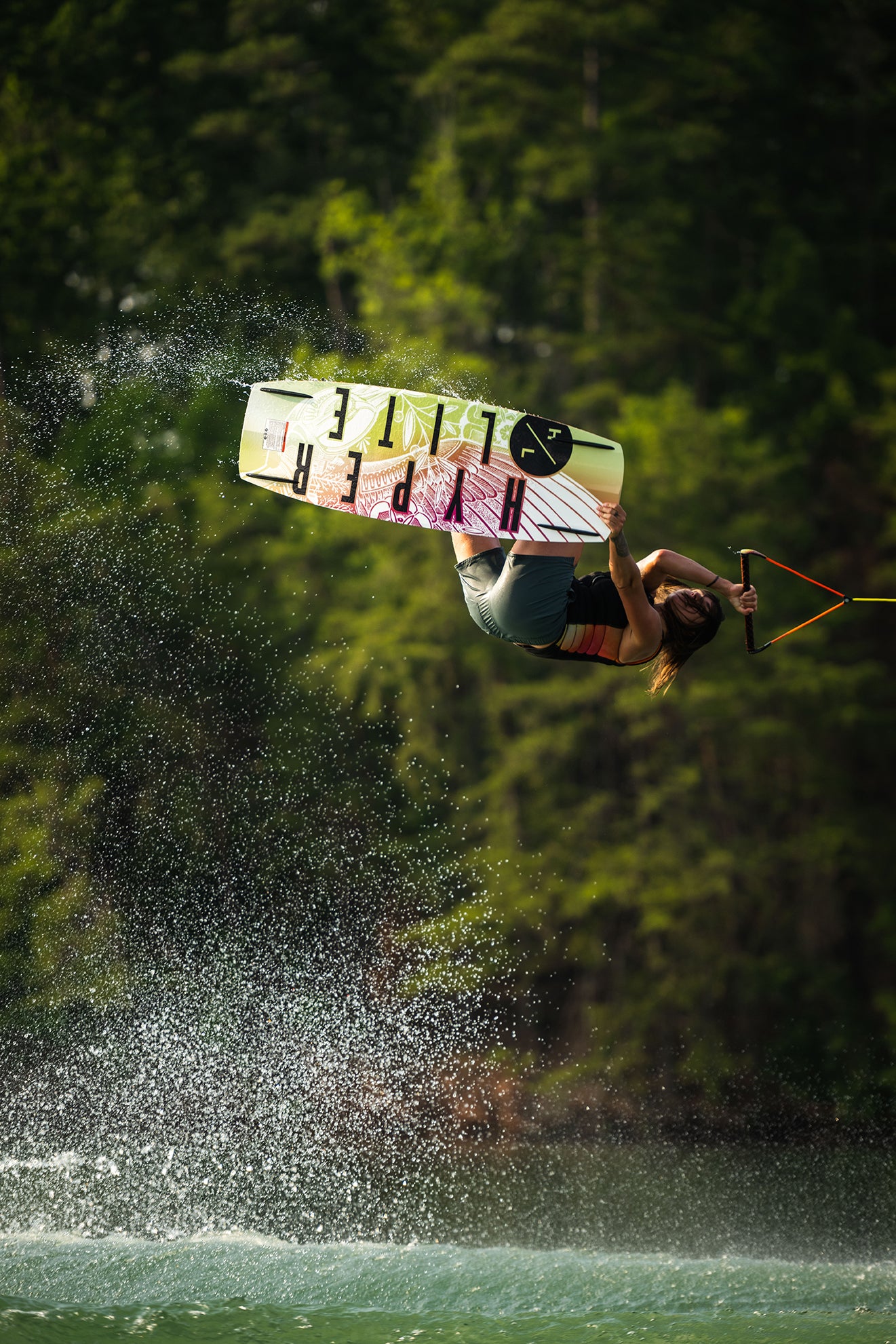 Person wakeboarding, performing an aerial trick over water, surrounded by trees.