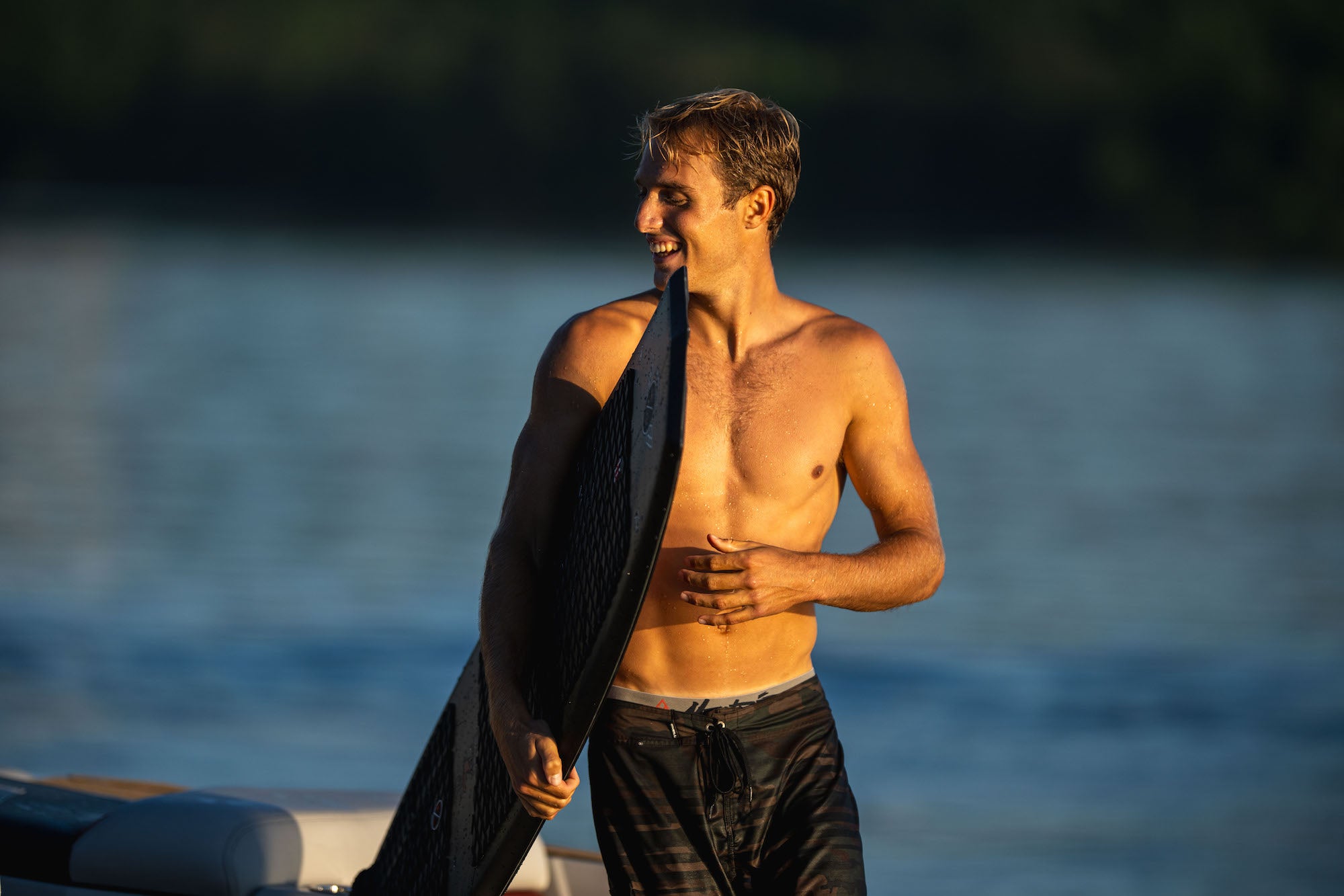 Shirtless man smiles while holding a wakesurf board on a boat at sunset.