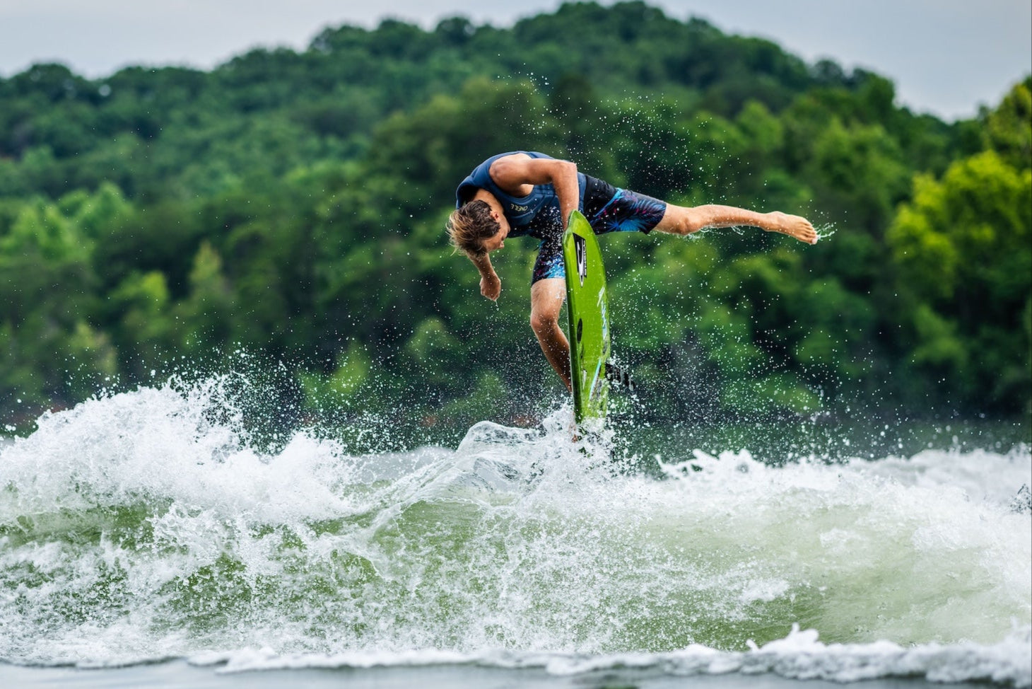 Wakesurfer twists into a trick with green board, water splashing around.