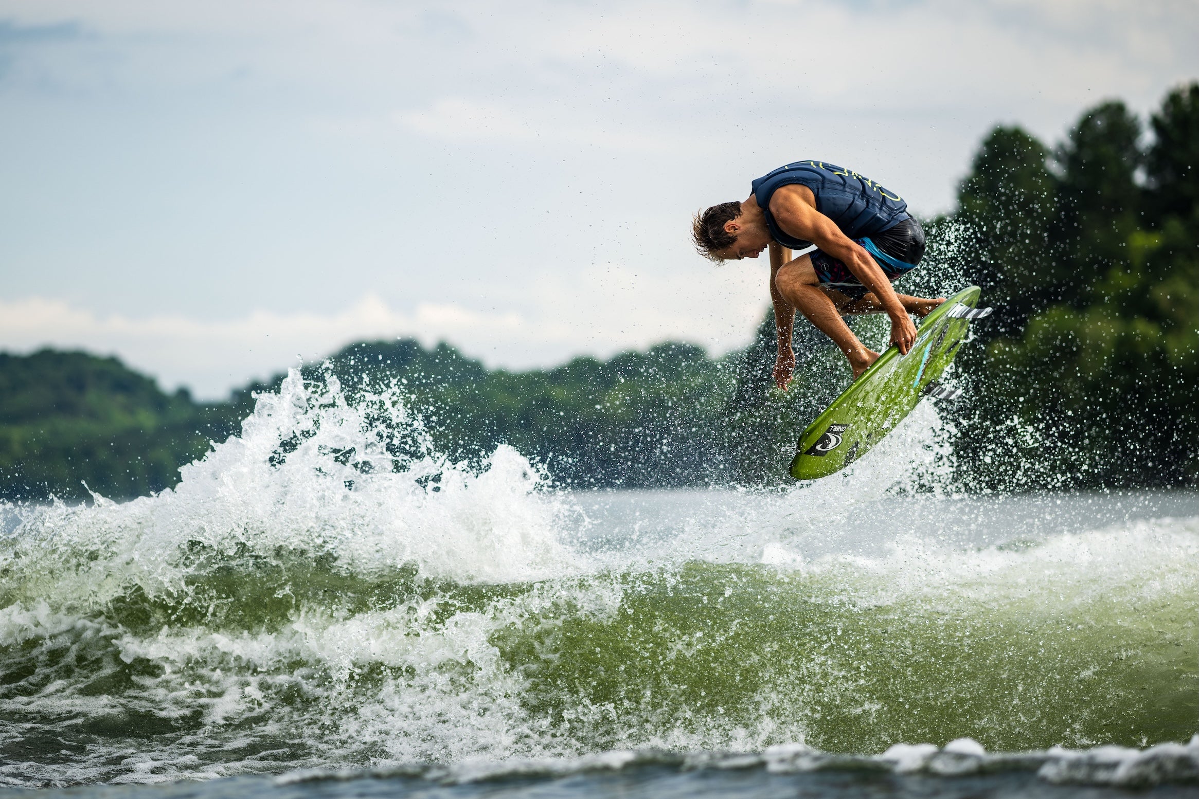Wakesurfer grabs bright green board midair while riding a breaking wake.